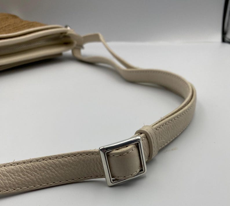 May include: A cream-colored shoulder bag strap made of textured leather with a silver-toned metal square buckle for length adjustment. The strap is attached to a tan-colored bag body that is partially visible in the upper left corner of the frame.