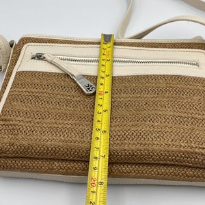 May include: A rectangular crossbody bag with a woven tan straw-like body and cream-colored leather trim. A yellow measuring tape placed vertically across the center shows the bag height is approximately 8 inches. It features a silver zipper.