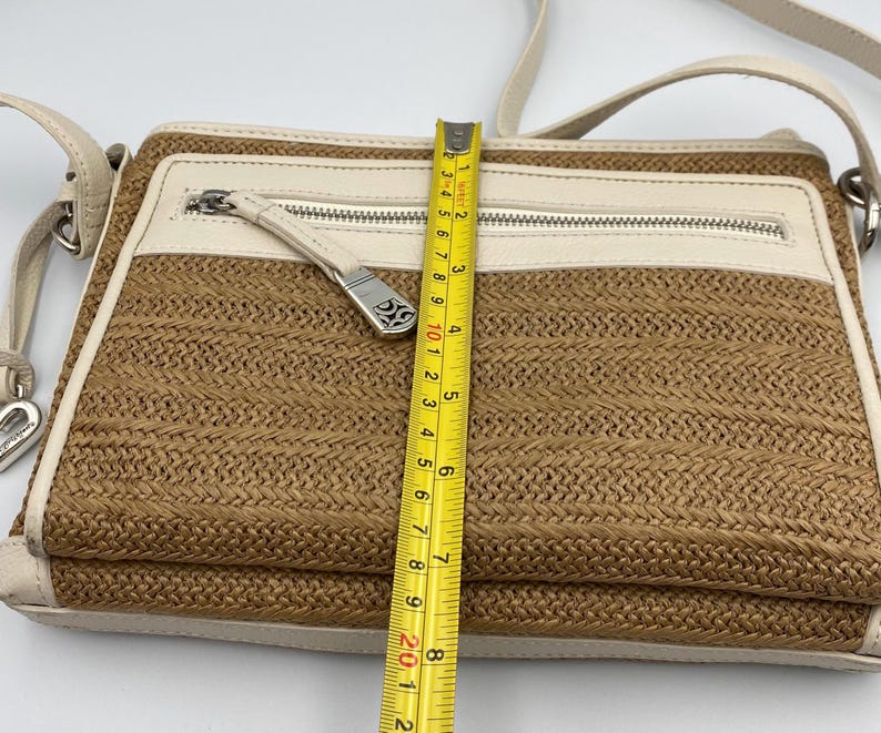 May include: A rectangular crossbody bag with a woven tan straw-like body and cream-colored leather trim. A yellow measuring tape placed vertically across the center shows the bag height is approximately 8 inches. It features a silver zipper.