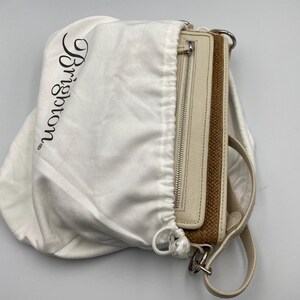 May include: A cream-colored leather handbag with a woven brown panel and a silver zipper, partially enclosed in a white fabric dust bag featuring the black printed text Brighton. The bag includes a matching cream leather strap and silver hardware.