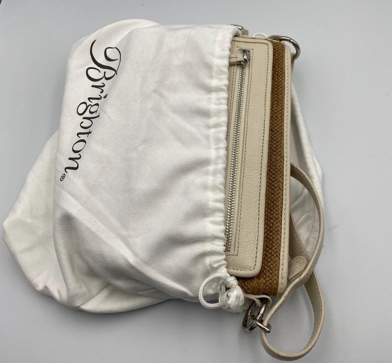 May include: A cream-colored leather handbag with a woven brown panel and a silver zipper, partially enclosed in a white fabric dust bag featuring the black printed text Brighton. The bag includes a matching cream leather strap and silver hardware.
