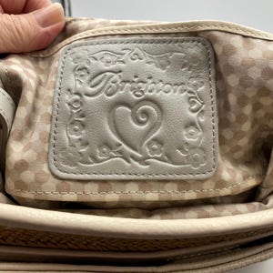 May include: A light beige leather interior patch featuring the embossed Brighton logo, a central heart, and a floral border. The patch is stitched onto a light beige fabric lining with a repeating white polka dot pattern inside a handbag.