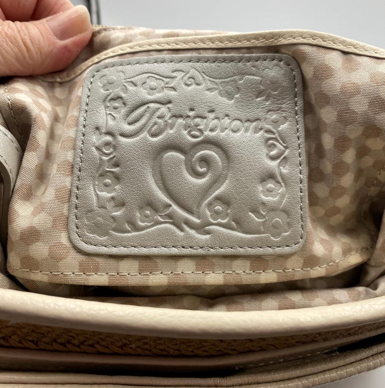 May include: A light beige leather interior patch featuring the embossed Brighton logo, a central heart, and a floral border. The patch is stitched onto a light beige fabric lining with a repeating white polka dot pattern inside a handbag.