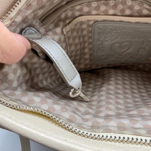 May include: The interior of a beige handbag featuring a light-colored fabric lining with a repeating pattern of small circles. A gray leather strap with a silver-toned metal clip is attached to the inside, alongside a gray leather patch embossed with the Brighton brand logo and a heart design.