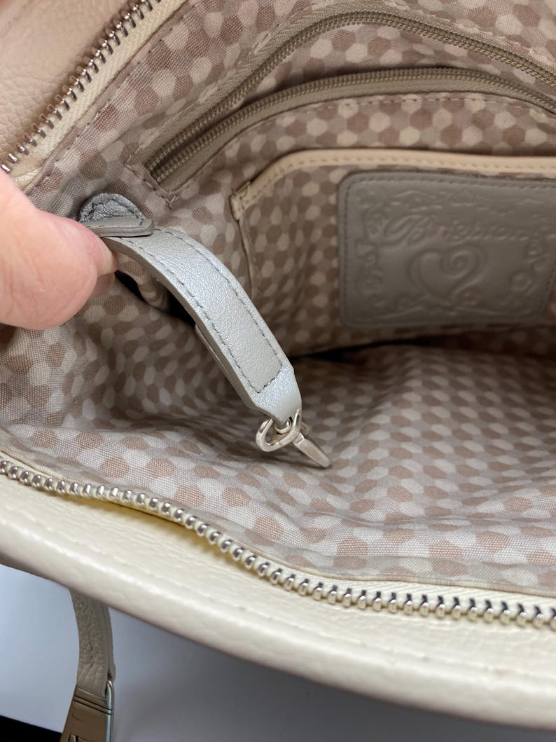 May include: The interior of a beige handbag featuring a light-colored fabric lining with a repeating pattern of small circles. A gray leather strap with a silver-toned metal clip is attached to the inside, alongside a gray leather patch embossed with the Brighton brand logo and a heart design.