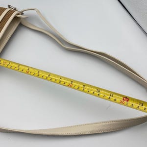 May include: A brown woven crossbody bag with cream-colored trim and a matching adjustable strap. A yellow measuring tape is placed across the bag, showing a width of approximately 14 inches. The bag features silver-toned metal hardware.
