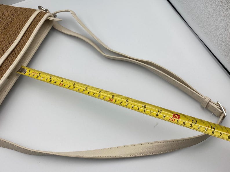 May include: A brown woven crossbody bag with cream-colored trim and a matching adjustable strap. A yellow measuring tape is placed across the bag, showing a width of approximately 14 inches. The bag features silver-toned metal hardware.