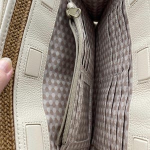 May include: An open cream-colored handbag interior featuring a central zippered pocket, multiple vertical card slots, and a clear identification window. The lining is a light fabric with a repeating geometric pattern, and the outer edge is woven tan straw.
