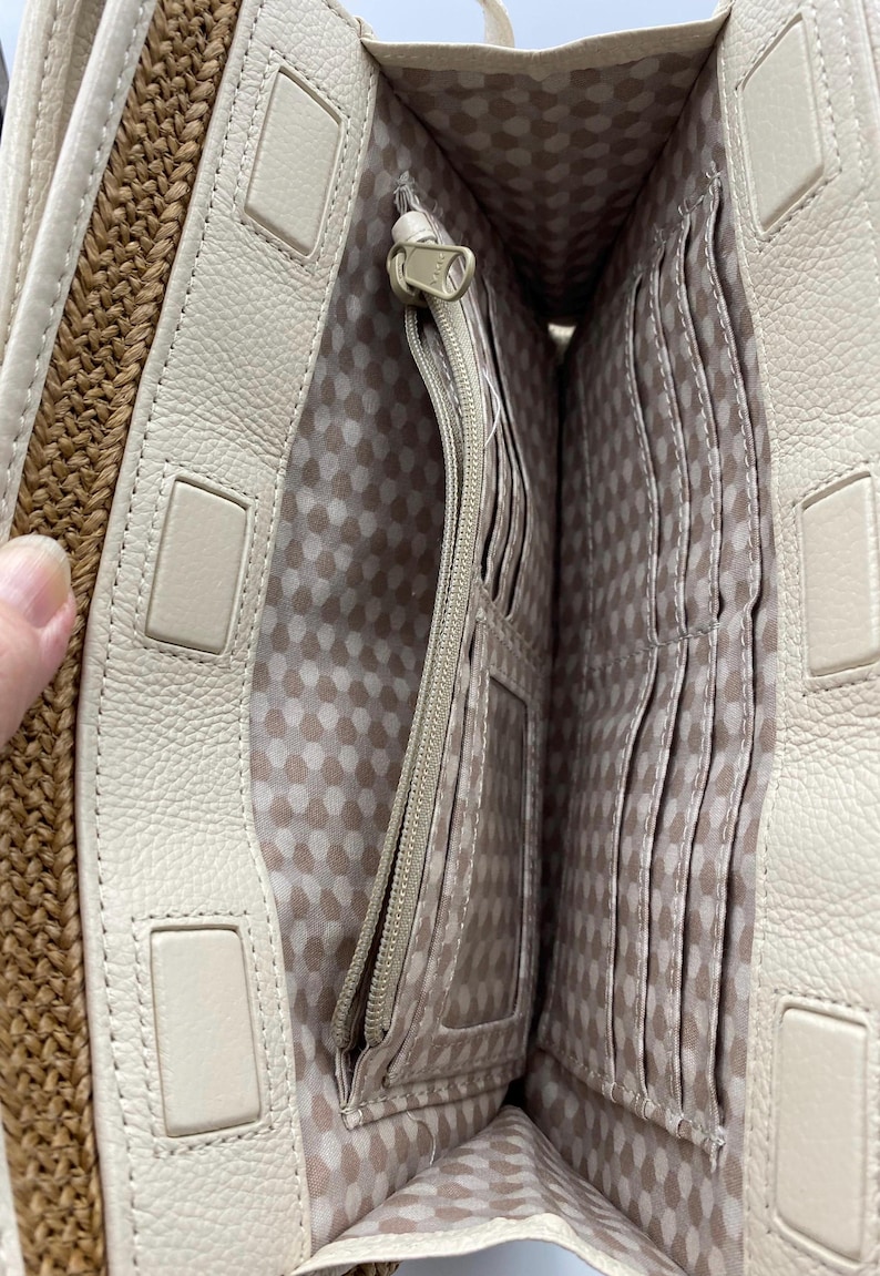 May include: An open cream-colored handbag interior featuring a central zippered pocket, multiple vertical card slots, and a clear identification window. The lining is a light fabric with a repeating geometric pattern, and the outer edge is woven tan straw.