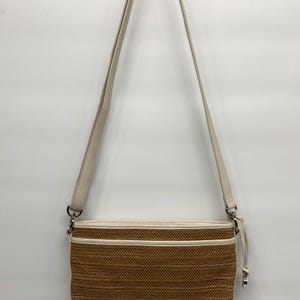 May include: A rectangular crossbody bag featuring a woven tan straw-like texture with cream-colored faux leather trim and piping. The bag includes an adjustable cream shoulder strap with a silver-toned metal buckle and a matching side zipper pull.