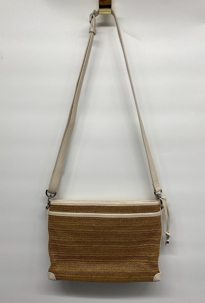 May include: A rectangular crossbody bag featuring a woven tan straw-like texture with cream-colored faux leather trim and piping. The bag includes an adjustable cream shoulder strap with a silver-toned metal buckle and a matching side zipper pull.