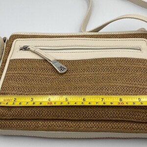 May include: A rectangular crossbody bag with a woven tan body and cream-colored leather trim. A yellow measuring tape across the front shows a width of approximately 10 inches. The bag features a zippered front pocket with a silver-toned metal pull.