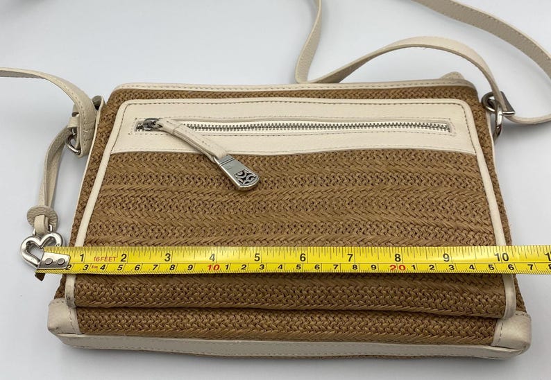 May include: A rectangular crossbody bag with a woven tan body and cream-colored leather trim. A yellow measuring tape across the front shows a width of approximately 10 inches. The bag features a zippered front pocket with a silver-toned metal pull.