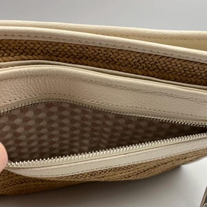 May include: A beige leather and woven straw clutch bag featuring a silver metal zipper closure. The interior is lined with a light-colored fabric patterned with a repeating hexagonal geometric design, visible through the open zippered compartment.