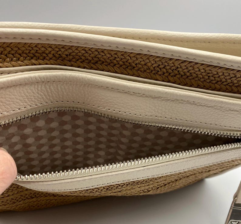 May include: A beige leather and woven straw clutch bag featuring a silver metal zipper closure. The interior is lined with a light-colored fabric patterned with a repeating hexagonal geometric design, visible through the open zippered compartment.