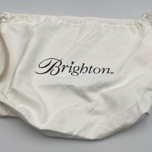 May include: A white fabric drawstring dust bag featuring the black text Brighton with a small heart above the letter i. The bag includes a light beige adjustable strap with a silver metal buckle attached to the side, designed for storage.