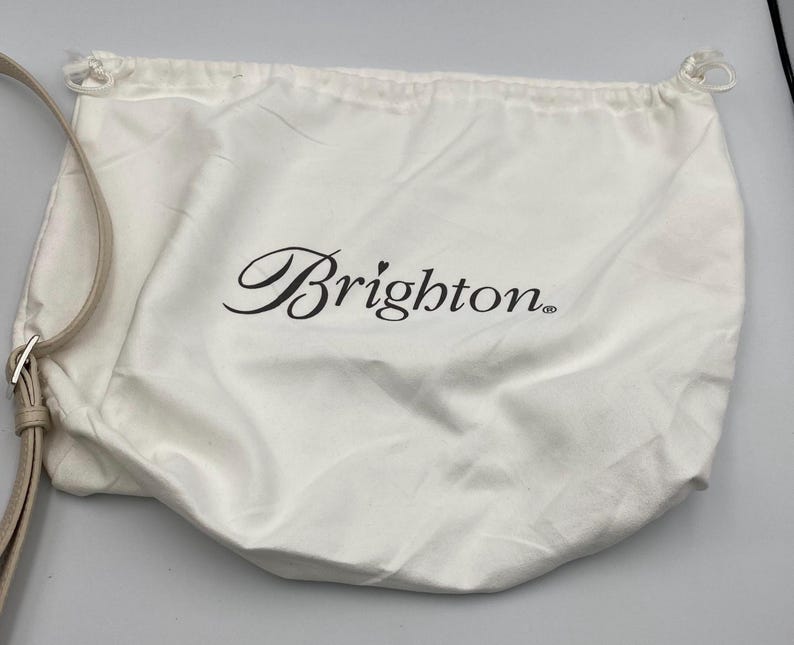 May include: A white fabric drawstring dust bag featuring the black text Brighton with a small heart above the letter i. The bag includes a light beige adjustable strap with a silver metal buckle attached to the side, designed for storage.