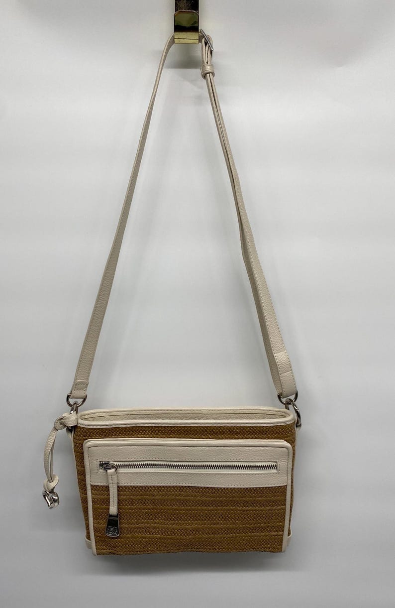 May include: A rectangular crossbody bag featuring a textured tan woven body with cream-colored leather trim. The bag includes an adjustable cream shoulder strap, a front zippered pocket with a silver-tone metal pull, and silver-tone hardware accents.