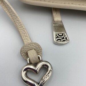 May include: A cream-colored textured leather handbag featuring a silver-tone metal heart-shaped charm with the word Brighton engraved on it. The charm is attached to a leather pull tab, and a separate silver-tone metal zipper pull with a cutout pattern.