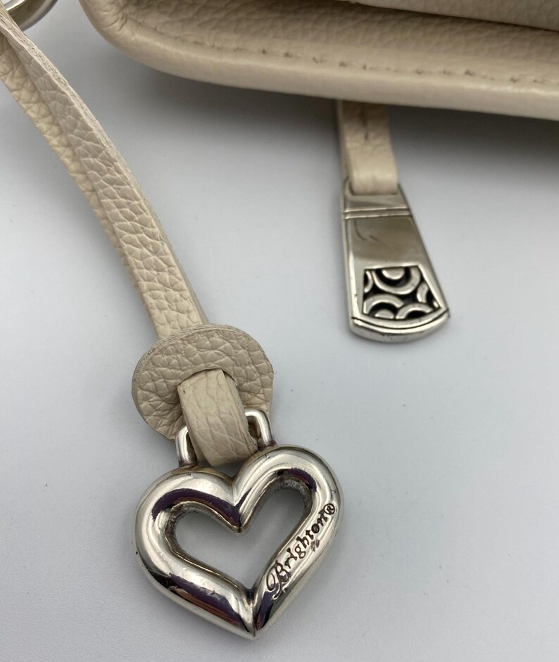 May include: A cream-colored textured leather handbag featuring a silver-tone metal heart-shaped charm with the word Brighton engraved on it. The charm is attached to a leather pull tab, and a separate silver-tone metal zipper pull with a cutout pattern.