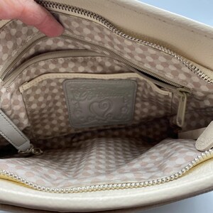 May include: The interior of a cream-colored leather handbag featuring a silver metal zipper closure and a light-colored fabric lining with a repeating geometric pattern. A rectangular leather patch embossed with the word Brighton and a heart icon.