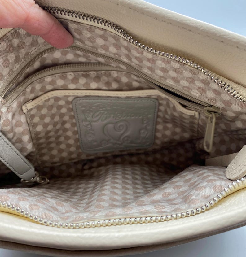 May include: The interior of a cream-colored leather handbag featuring a silver metal zipper closure and a light-colored fabric lining with a repeating geometric pattern. A rectangular leather patch embossed with the word Brighton and a heart icon.