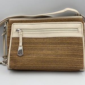 May include: A rectangular crossbody bag featuring a woven tan straw body with cream-colored leather trim. The bag includes a front zippered pocket with a silver-toned metal pull, a matching cream leather shoulder strap, and silver-toned hardware.