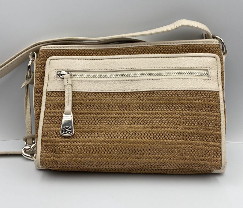 May include: A rectangular crossbody bag featuring a woven tan straw body with cream-colored leather trim. The bag includes a front zippered pocket with a silver-toned metal pull, a matching cream leather shoulder strap, and silver-toned hardware.
