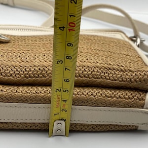 May include: A beige and tan woven crossbody bag with white leather trim and a silver zipper pull. A yellow measuring tape is positioned vertically against the side, showing a height measurement of approximately 4.25 inches for the bag's profile.