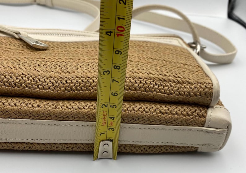 May include: A beige and tan woven crossbody bag with white leather trim and a silver zipper pull. A yellow measuring tape is positioned vertically against the side, showing a height measurement of approximately 4.25 inches for the bag's profile.