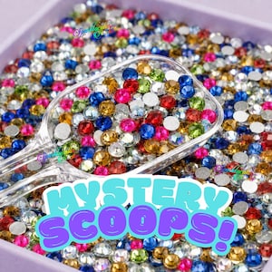 May include: A clear plastic scoop filled with a vibrant assortment of multi-coloured round rhinestones in shades of blue, red, green, yellow, and clear. The text MYSTERY SCOOPS! is displayed in large, bold, light blue and purple letters.