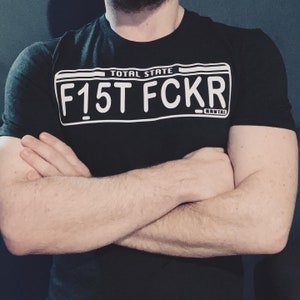 May include: A black t-shirt featuring a white license plate graphic that reads "TOTAL STATE F15T FCKR BRUTAL".