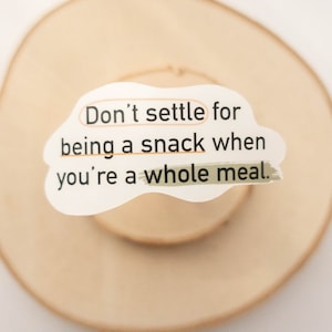May include: A white paper cutout with the text "Don't settle for being a snack when you're a whole meal." The text is highlighted with orange and green lines. The cutout is placed on a light brown wooden surface.