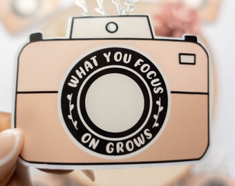 Puede incluir: Una pegatina de cámara rosa y blanca con el texto "What you focus on grows" en un círculo negro. La cámara tiene un diseño de flores blancas en la parte superior.