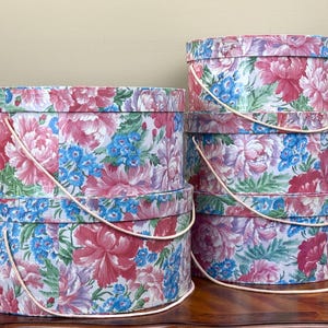 May include: A stack of floral hat boxes in various sizes. The boxes are cylindrical with a white background and a pattern of pink, blue, and purple flowers. Each box has a white rope handle.
