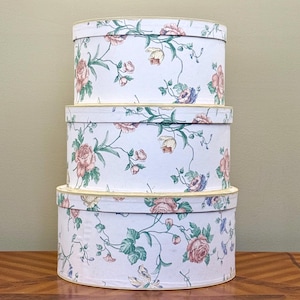 May include: Three oval-shaped decorative boxes stacked on top of each other. The boxes are white with a floral pattern of pink roses, blue and yellow flowers, and green leaves. The edges of the boxes are trimmed with a gold color.