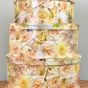 May include: Three stacked, round hat boxes with floral patterns. The boxes feature a repeating design of yellow and peach roses and other flowers. Each box has a white handle.