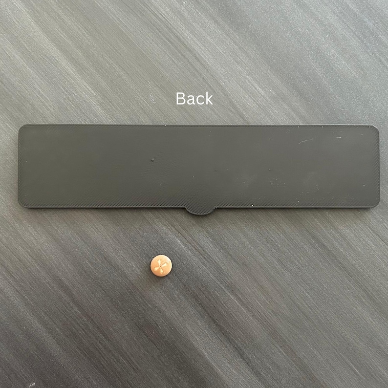 Puede incluir: Un componente rectangular de pl&aacute;stico negro con una peque&ntilde;a pesta&ntilde;a semicircular en el borde inferior, etiquetado con la palabra Back en texto blanco, colocado sobre un bot&oacute;n peque&ntilde;o, redondo y de color naranja claro con cuatro agujeros.