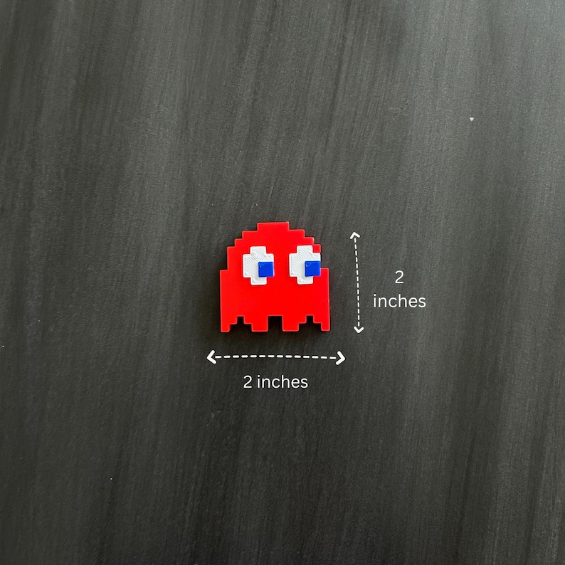 May include: Red pixelated ghost character with blue eyes and a white outline. The ghost is 2 inches tall and 2 inches wide.