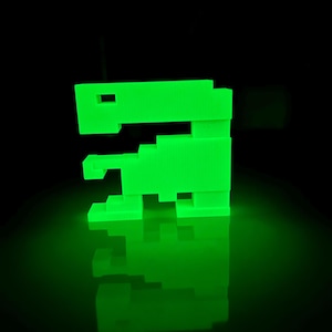 May include: A green glow-in-the-dark plastic toy shaped like a pixelated alien spaceship. The toy is sitting on a black surface and reflecting light.