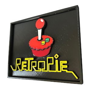 May include: A black framed sign featuring a red joystick and the word "RetroPie" in yellow. The joystick has a red ball, white stick, and a red base with yellow and green buttons. The sign is a 3D printed design.