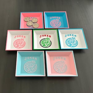 May include: Seven square trays with raised edges, each featuring the word JOKER and a circular graphic design. The trays come in various color combinations including pink, blue, white, and green. One tray contains four silver-colored coins.