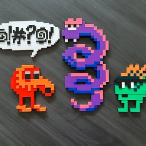 May include: A set of four pixel art figures, including a purple snake, a green figure with a black eye mask and orange hair, an orange figure with a beak, and a white speech bubble with the text "@!#?@!"