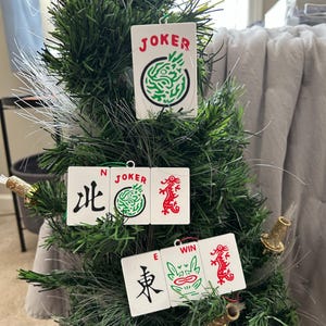 May include: White Mahjong tile ornaments with red and green designs hang on a green Christmas tree. The ornaments feature Chinese characters, dragons, and the word "JOKER". The tree is decorated with fiber optic lights.