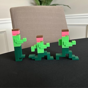 May include: Three pixelated figures in a running pose. The figures are constructed from green, pink, and brown blocks. The figures are on a black surface, with a neutral-colored chair and wall in the background.