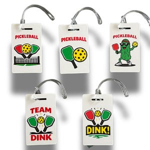 May include: White rectangular luggage tags with silver loops, featuring various pickleball-themed graphics. Designs include paddles, balls, nets, and the word "PICKLEBALL." One tag shows a pickle wearing sunglasses. The words "TEAM DINK" and "DINK!" are also displayed.