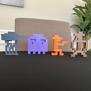 May include: Four colorful, pixelated figures from classic video games are displayed on a black surface. The figures include a gray AT-AT walker, a purple ghost, an orange character, and a brown cowboy.
