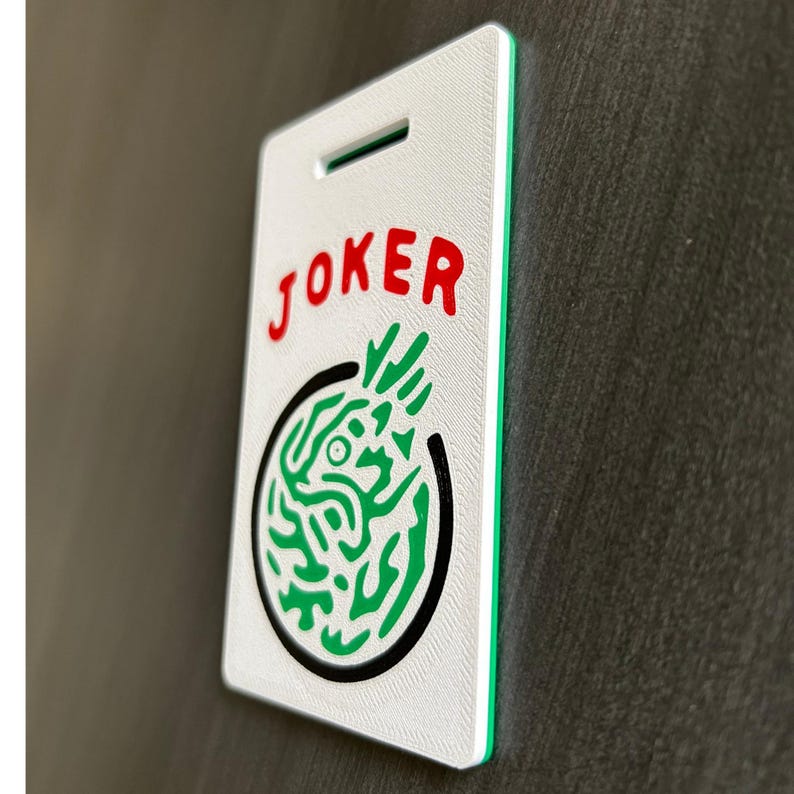 May include: A white rectangular tag with the word "JOKER" in red letters at the top. Below the word is a green and black circular design. The tag has a green border and a slot at the top.