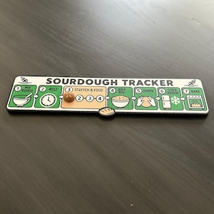 Puede incluir: Un tablero rectangular de seguimiento de masa madre con fondo blanco y detalles en verde. Incluye siete pasos numerados para hacer pan: mezclar, reposar, estirar y plegar, fermentar, dar forma, enfriar y hornear, con un peque&ntilde;o marcador marr&oacute;n.