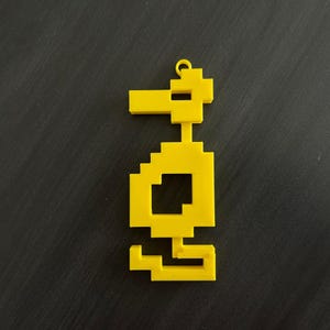 May include: A bright yellow, pixelated duck-shaped charm. The charm has a loop at the top for hanging and a square body. The duck is made of a solid, smooth material and is set against a dark background.